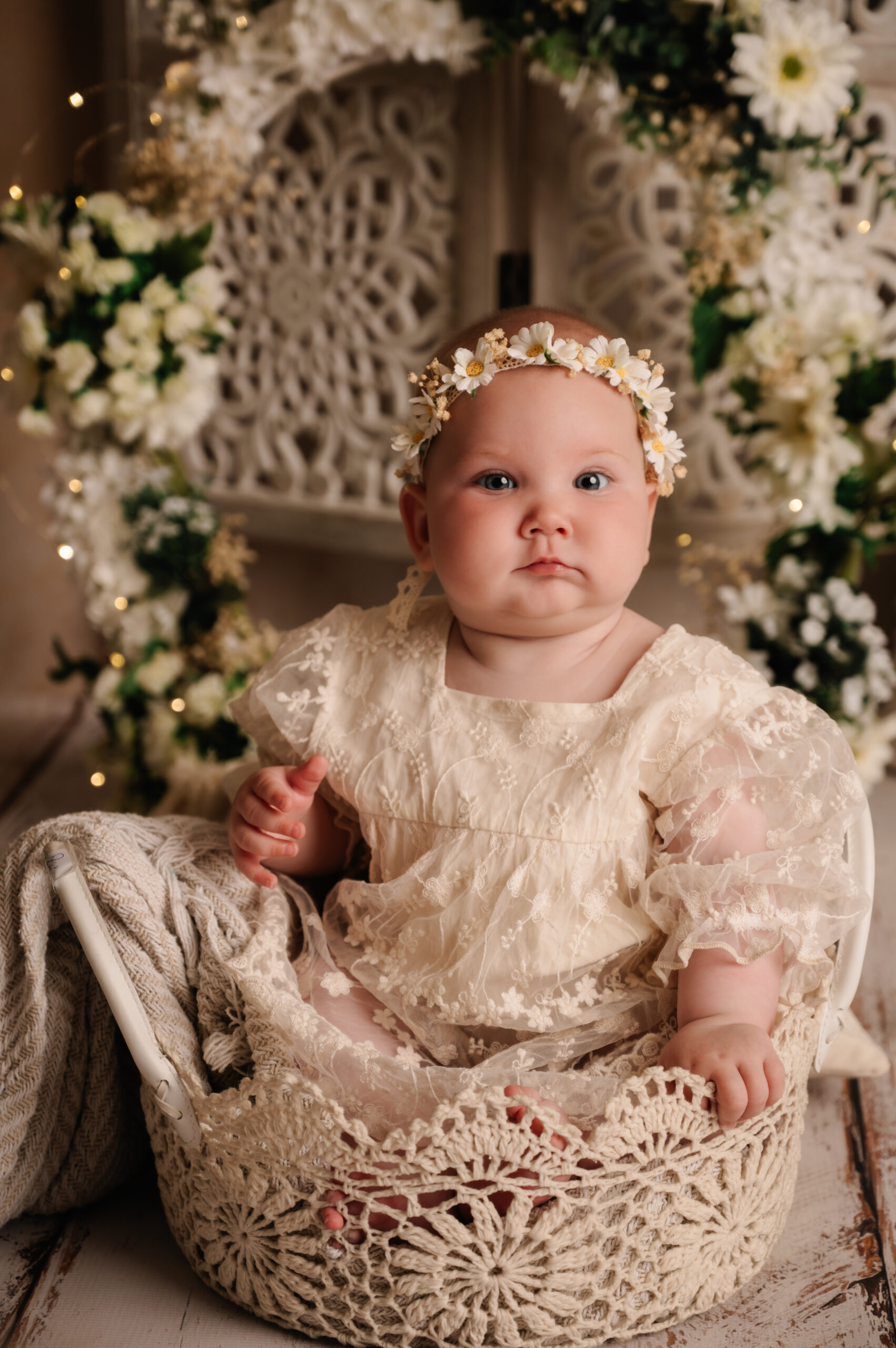 Newborn Fotoshoot Almere – Baby Portret in Kanten Outfit | Rebellens Fotografie Baby in kanten outfit en bloemendiadeem zit in een gehaakt mandje voor een bloemrijke achtergrond – newborn fotoshoot in studio Almere door Rebellens Fotografie.
