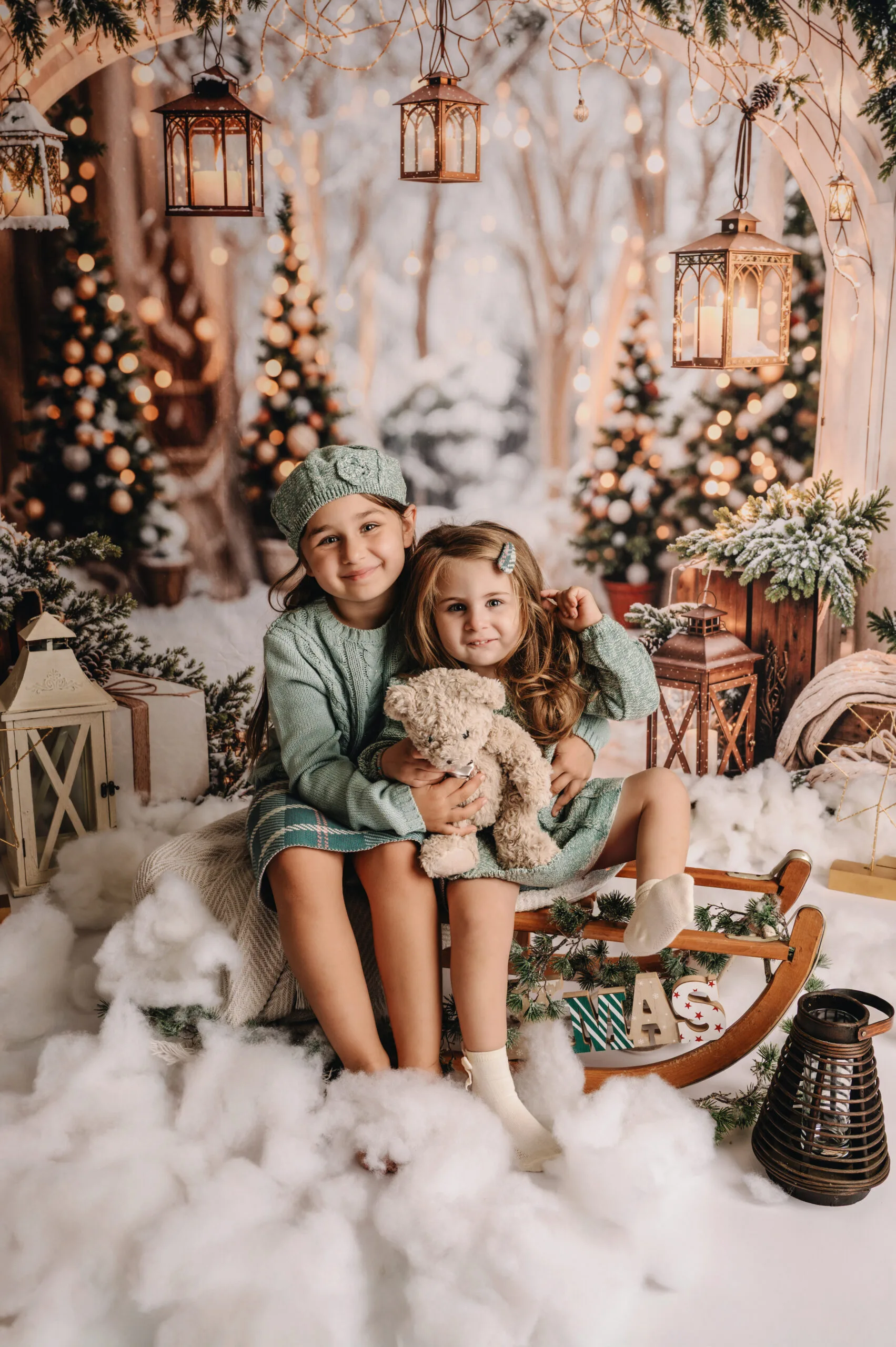 Twee jonge meisjes in mintgroene winteroutfits op een slee in een sfeervol kerstdecor met lichtjes, lantaarns en een knuffelbeer