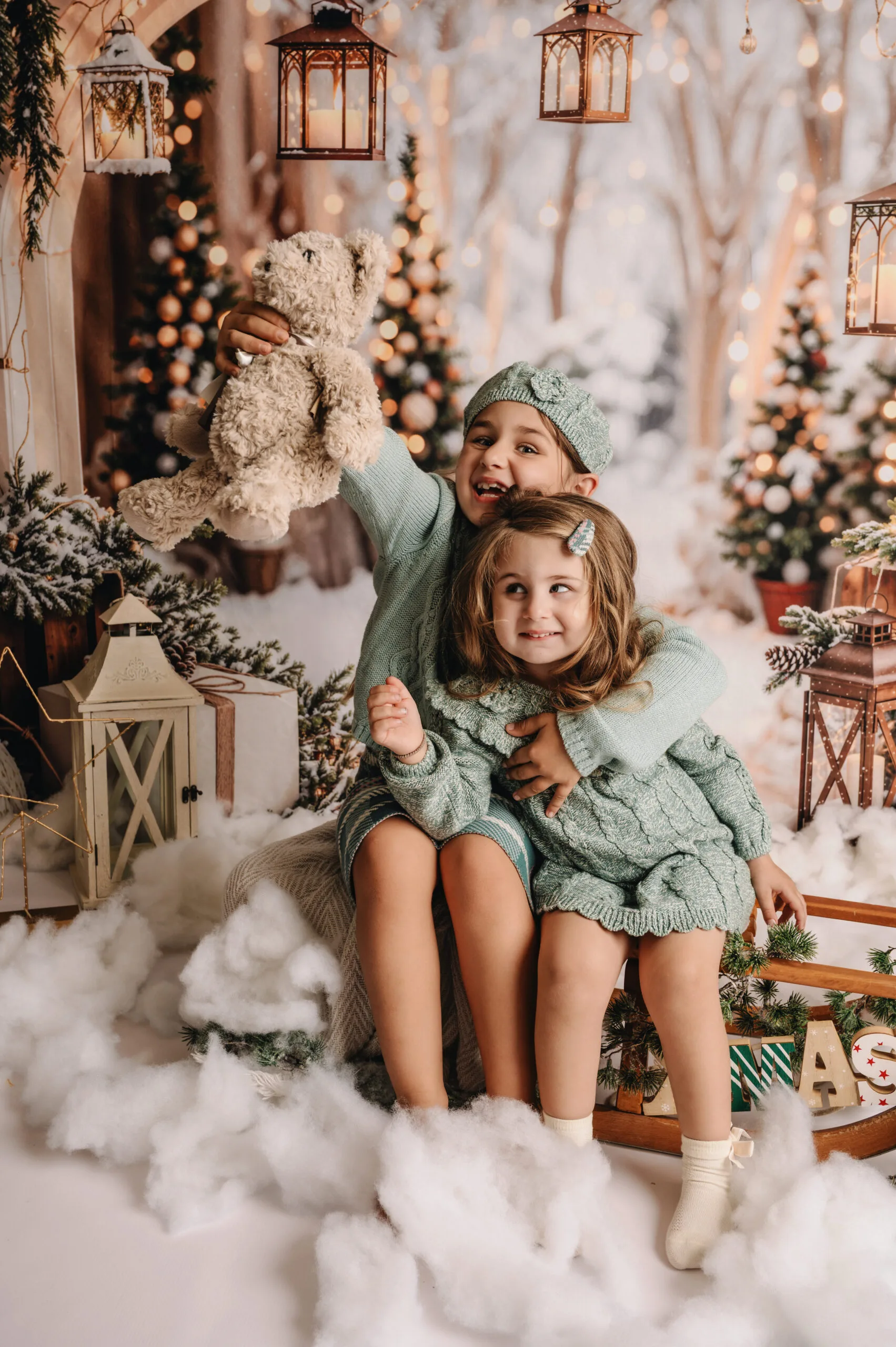 Twee jonge meisjes in mintgroene winteroutfits op een slee in een sfeervol kerstdecor met lichtjes, lantaarns en een knuffelbeer