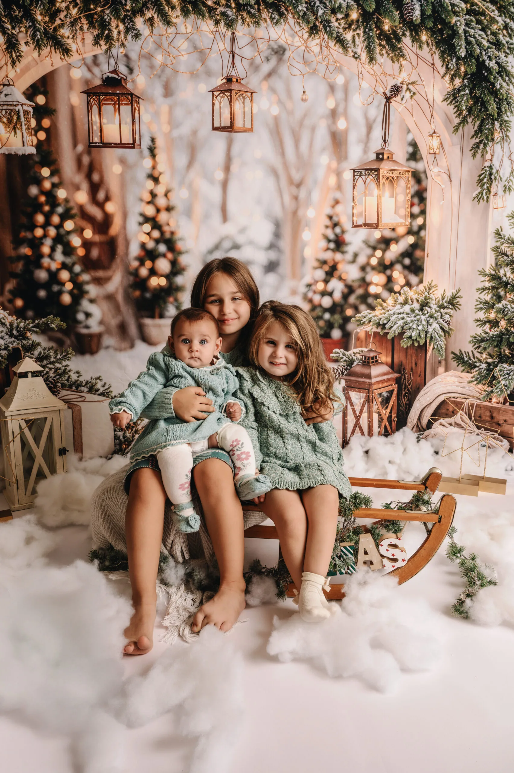 Twee jonge meisjes in mintgroene winteroutfits op een slee in een sfeervol kerstdecor met lichtjes, lantaarns en een knuffelbeer