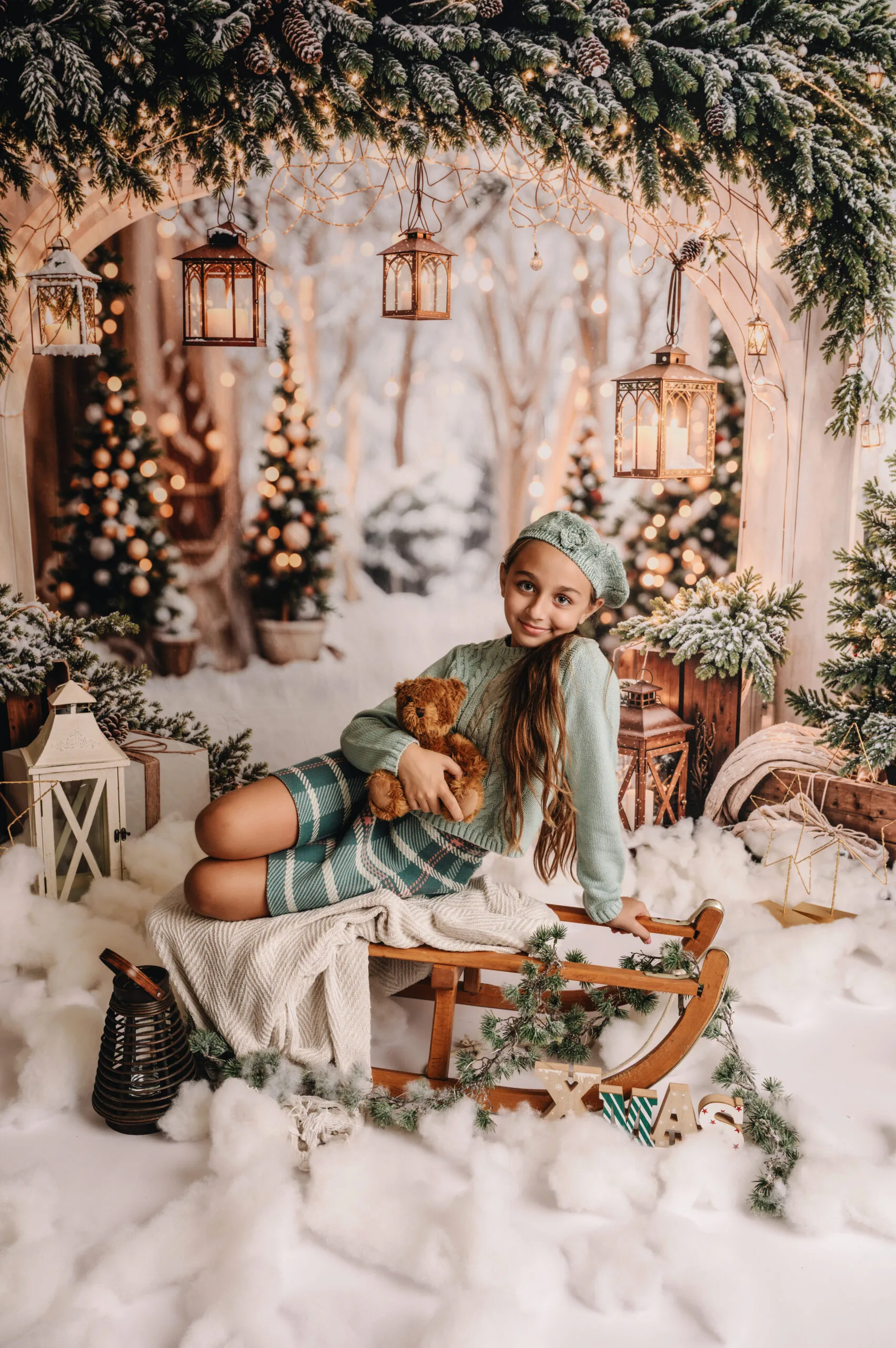 Twee jonge meisjes in mintgroene winteroutfits op een slee in een sfeervol kerstdecor met lichtjes, lantaarns en een knuffelbeer