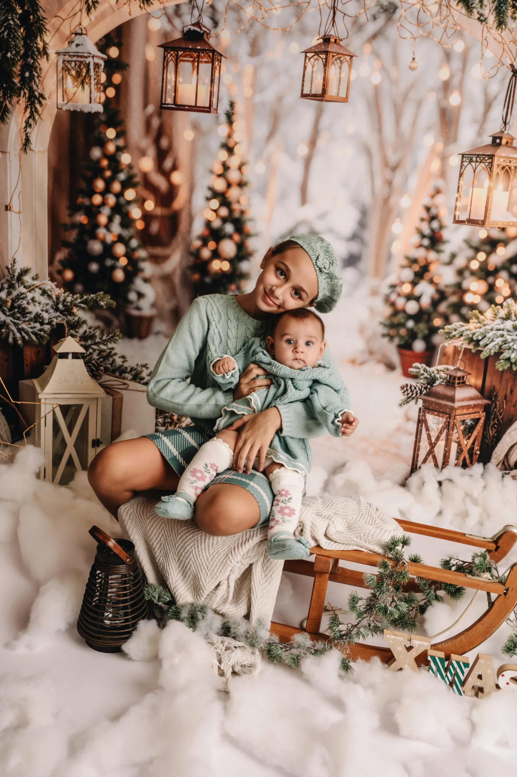 Twee jonge meisjes in mintgroene winteroutfits op een slee in een sfeervol kerstdecor met lichtjes, lantaarns en een knuffelbeer