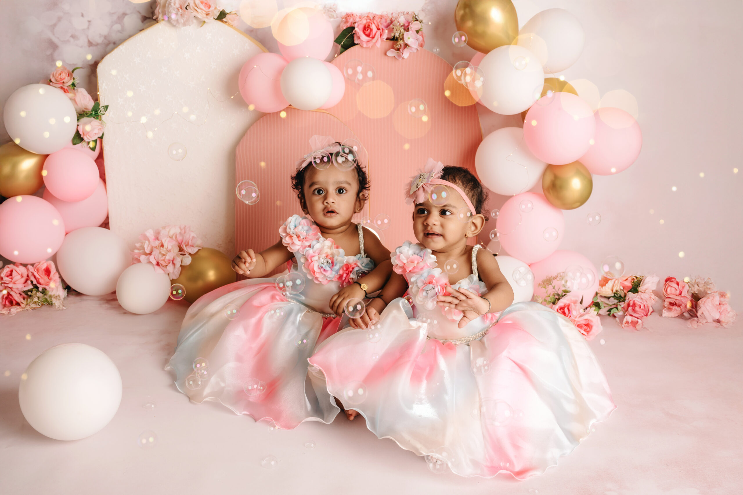 First Birthday Cake Smash Twins Two baby girls in matching pink dresses during a first birthday cake smash photoshoot with balloons, flowers, and a cake