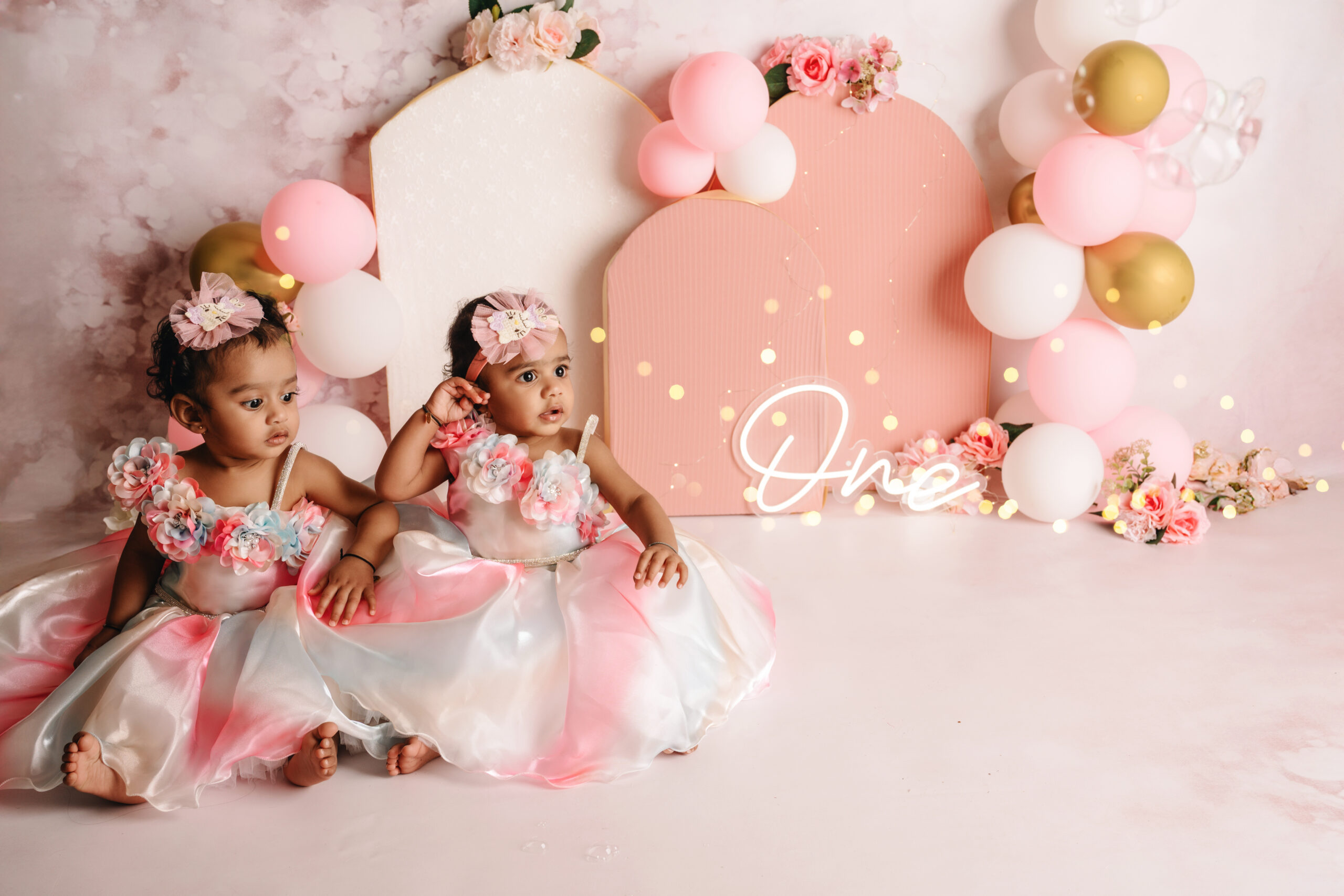 First Birthday Cake Smash Twins Two baby girls in matching pink dresses during a first birthday cake smash photoshoot with balloons, flowers, and a cake