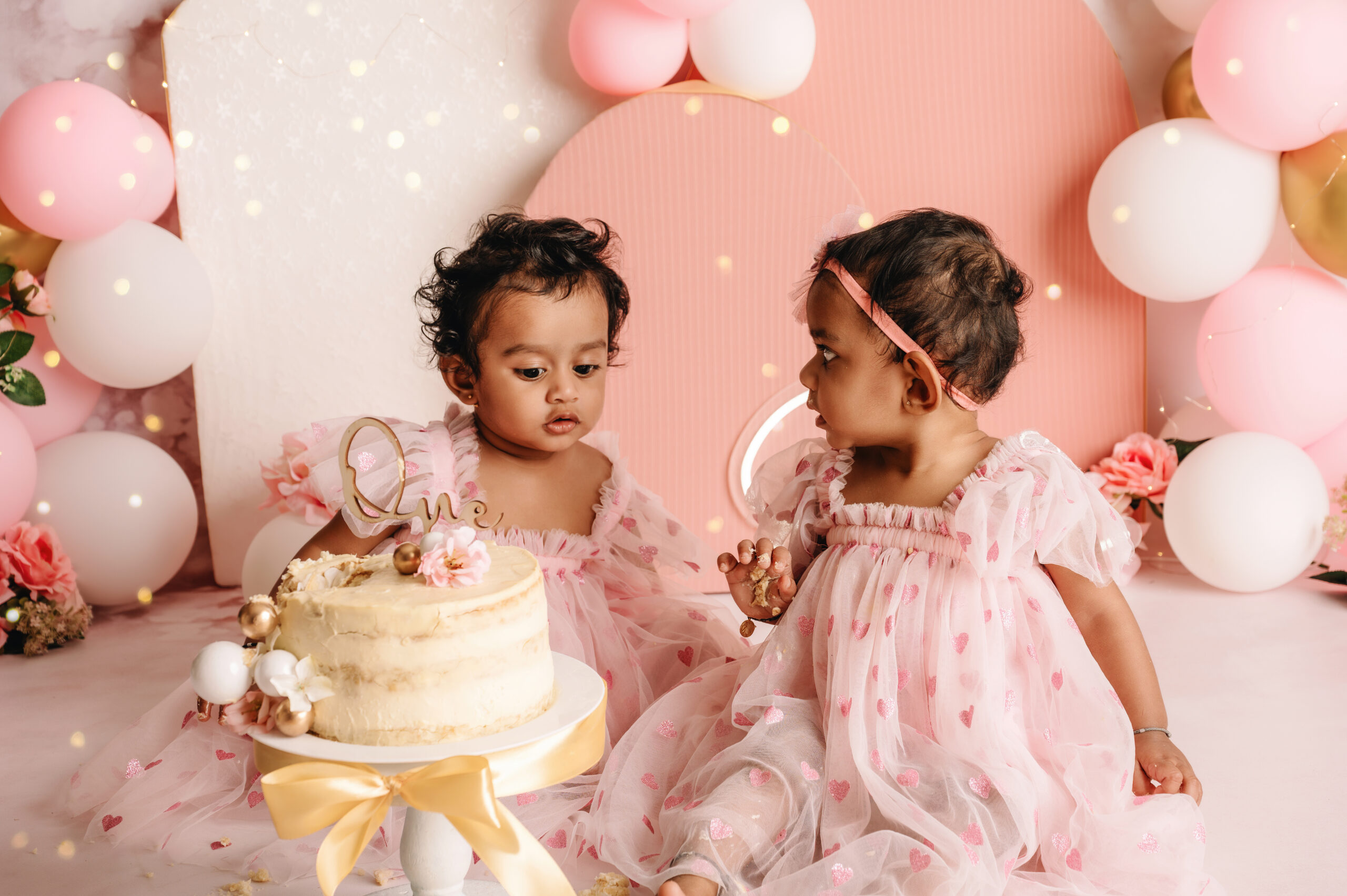 First Birthday Cake Smash Twins Two baby girls in matching pink dresses during a first birthday cake smash photoshoot with balloons, flowers, and a cake