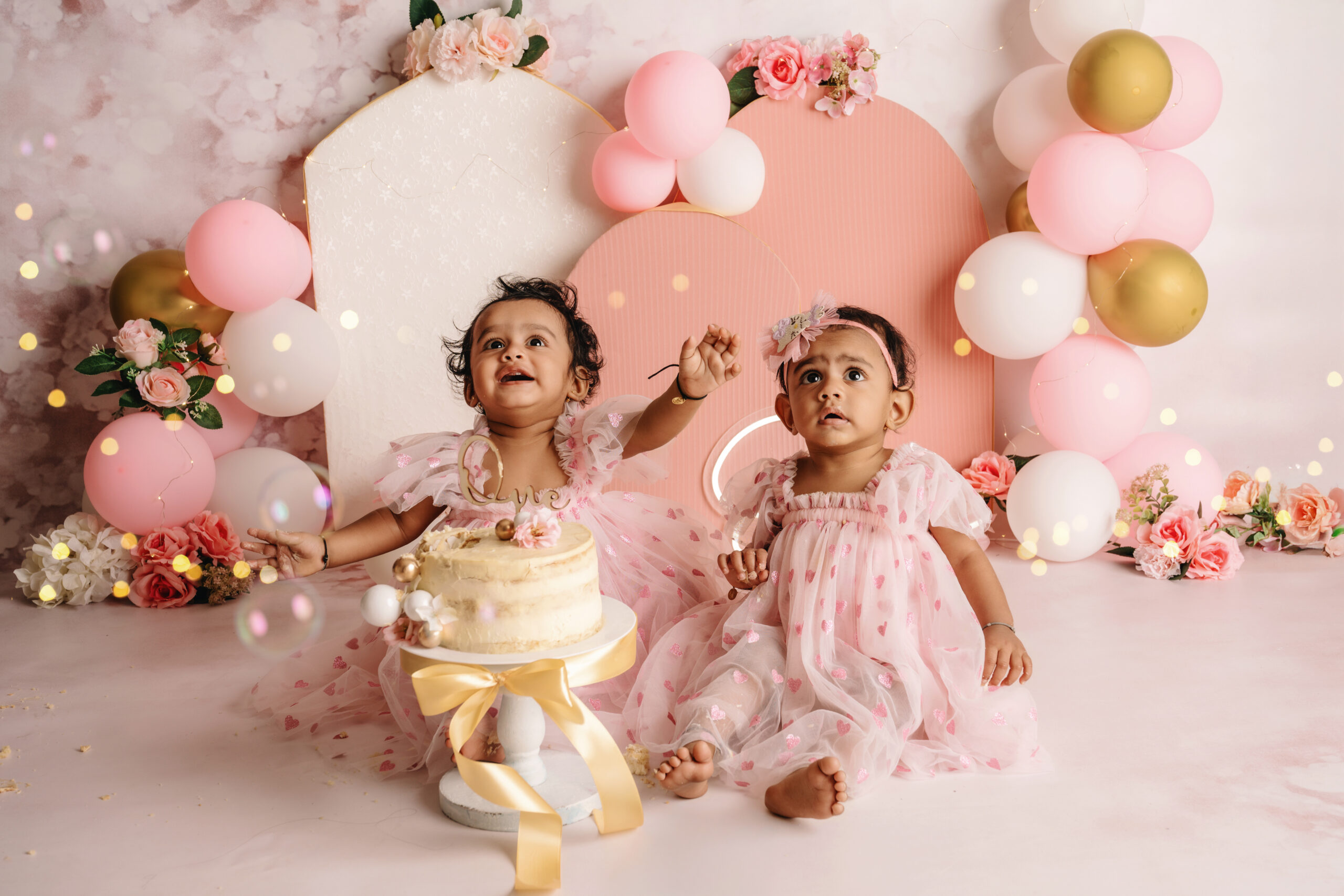 First Birthday Cake Smash Twins Two baby girls in matching pink dresses during a first birthday cake smash photoshoot with balloons, flowers, and a cake
