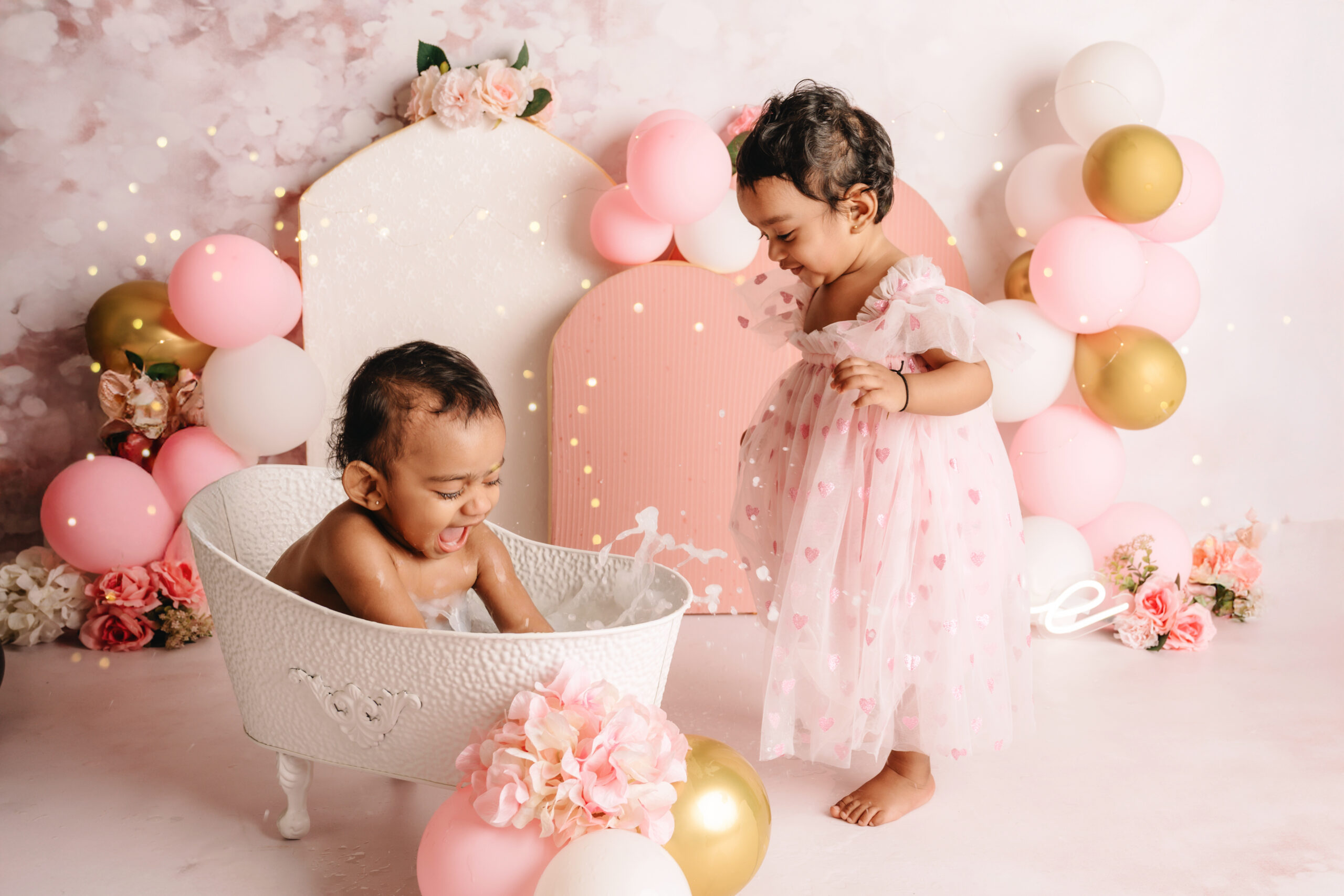 First Birthday Cake Smash Twins Two baby girls in matching pink dresses during a first birthday cake smash photoshoot with balloons, flowers, and a cake