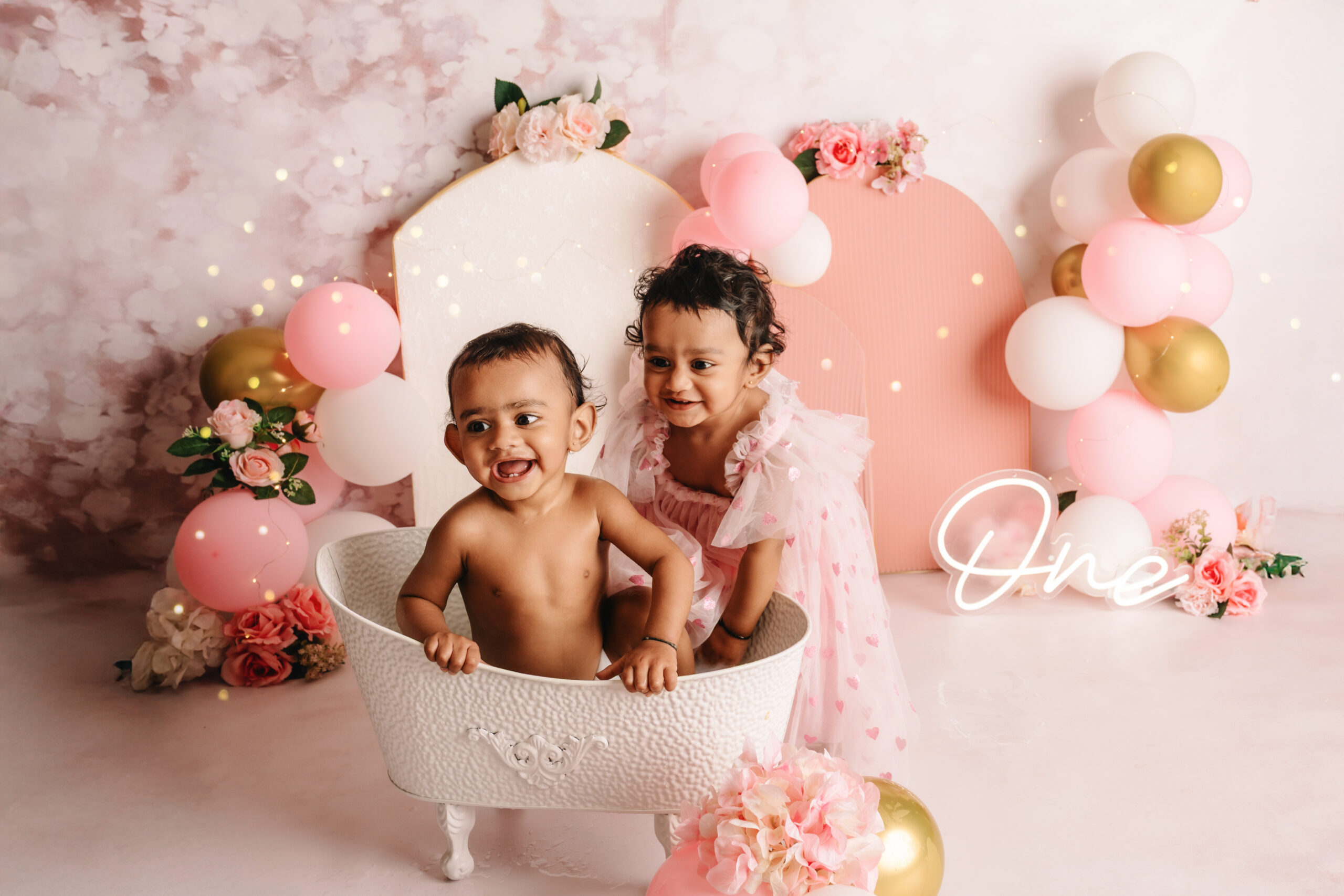 First Birthday Cake Smash Twins Two baby girls in matching pink dresses during a first birthday cake smash photoshoot with balloons, flowers, and a cake